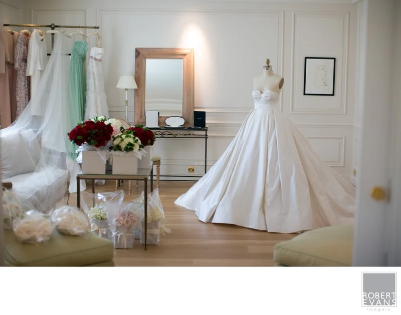 A wedding dress hanging in a softly lit room beside flowers and a mirror.