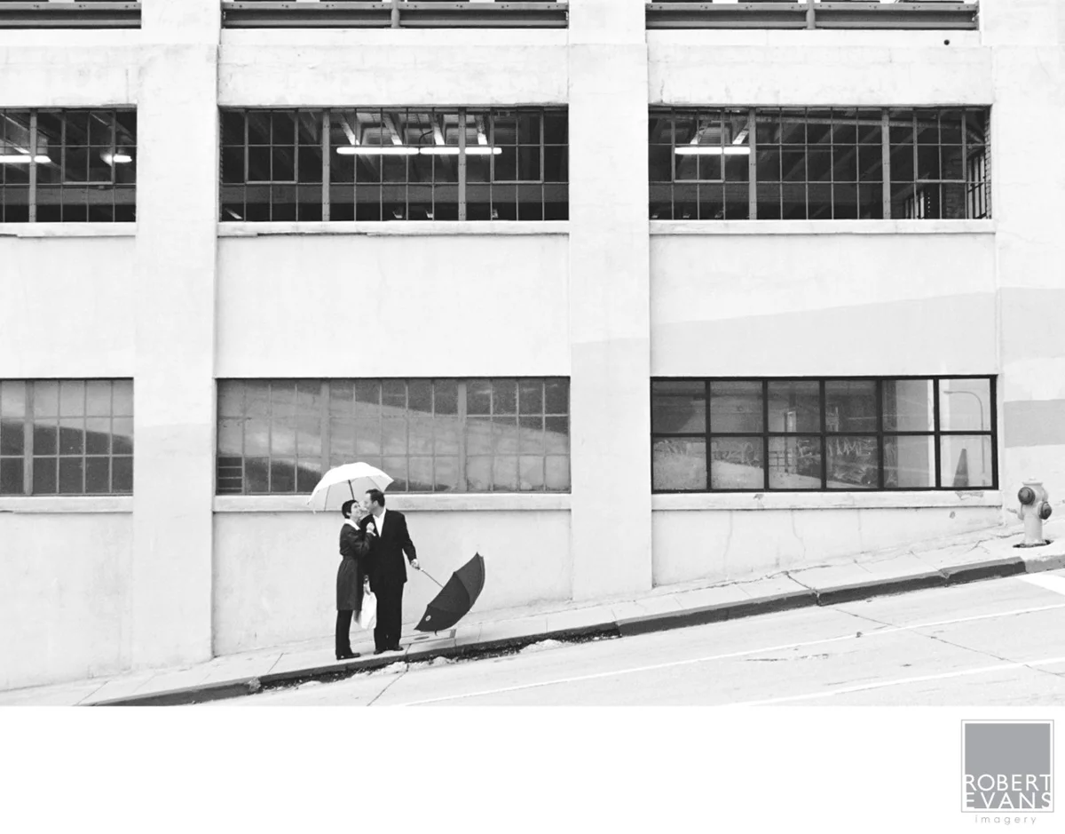 A couple stands beside a sunlit wall in a black-and-white city portrait.