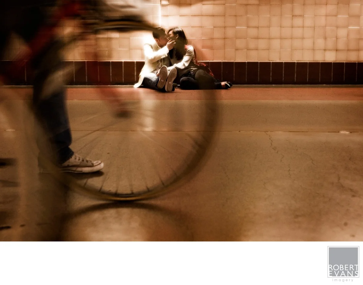 A couple kisses beside a tiled wall as a bicycle passes in the foreground.