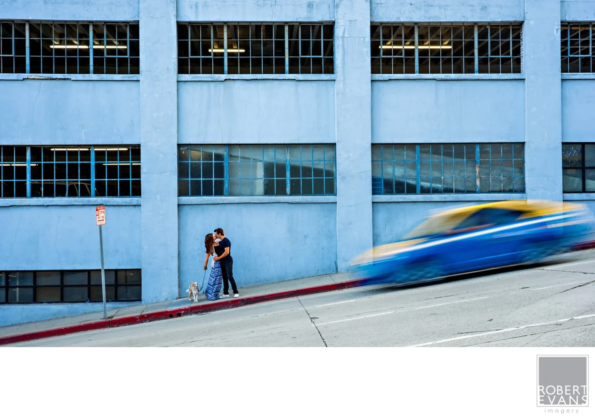 A couple stands against a blue building as a vehicle moves past.