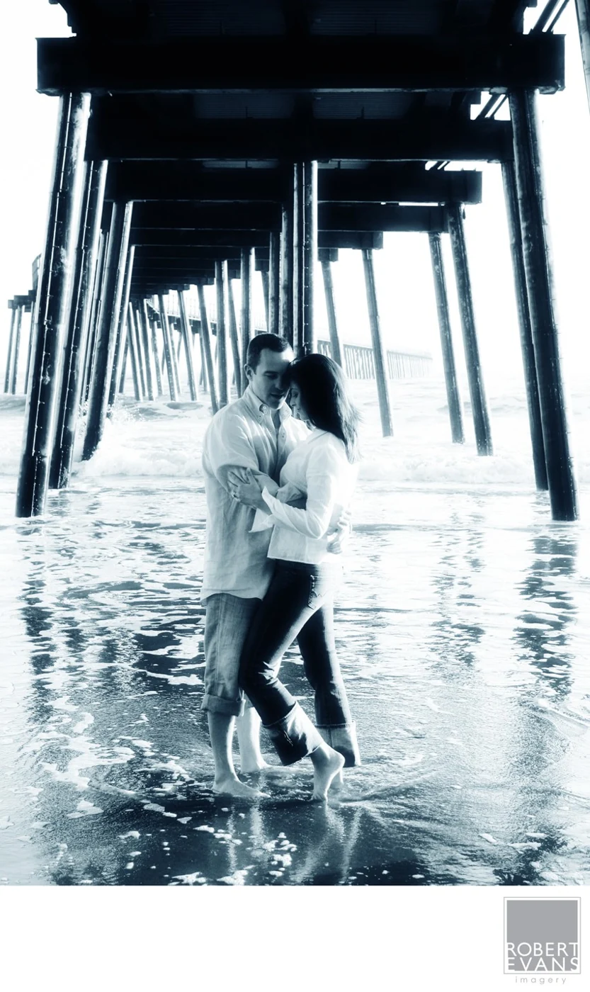 A couple embracing in shallow water beneath the repeating supports of a pier.