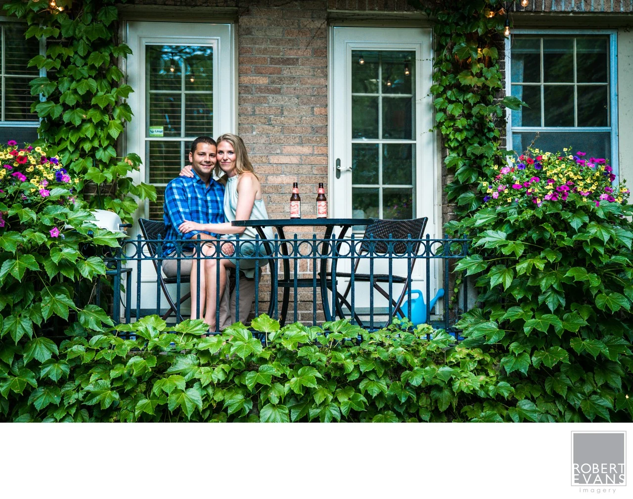 A couple together on a small balcony framed by green climbing plants and flowers.