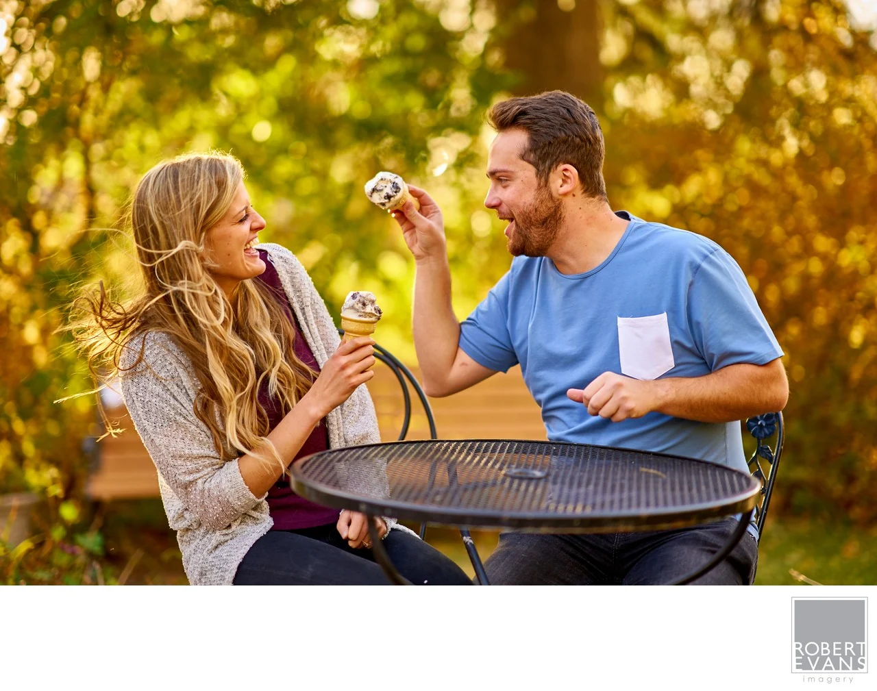A couple blowing bubbles toward each other across an outdoor table in autumn.