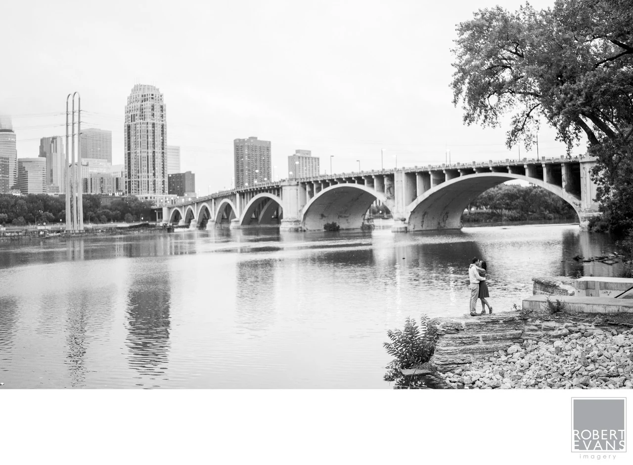 A black-and-white portrait of a couple beside a river with a stone bridge and city skyline beyond.