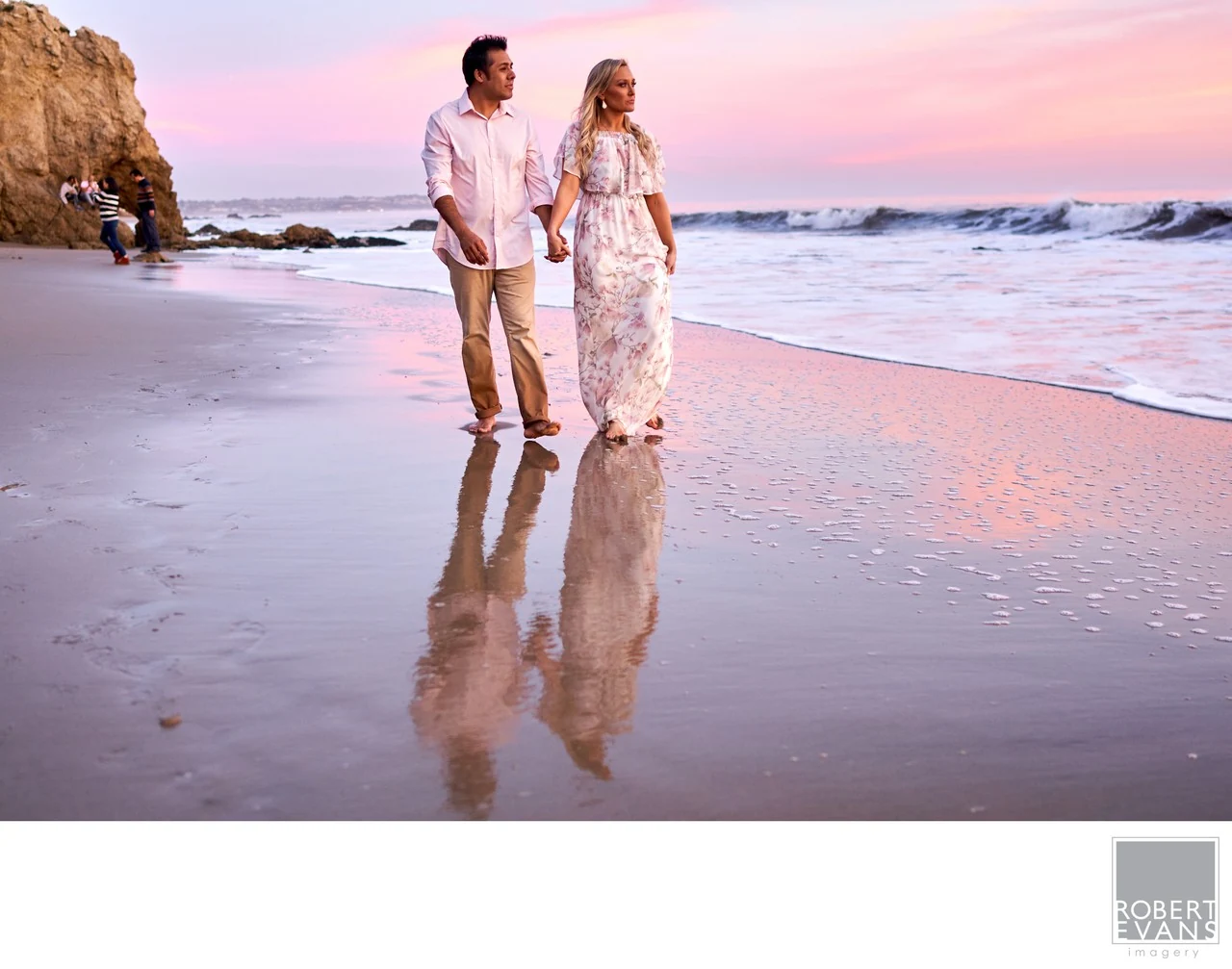 A couple walking hand in hand along a beach beneath a pink evening sky.