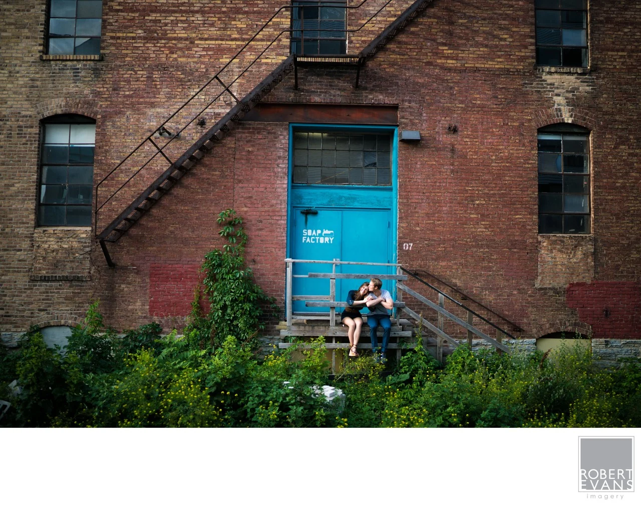A couple sitting on the steps beneath a blue door in a brick building.