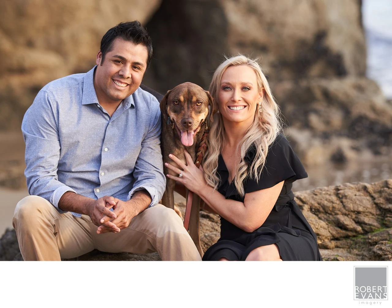A couple sitting close together with their dog beside a rocky shoreline.