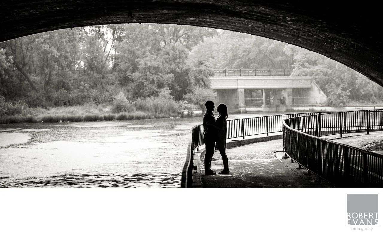 A black-and-white portrait of a couple by the water beneath the arch of a bridge.