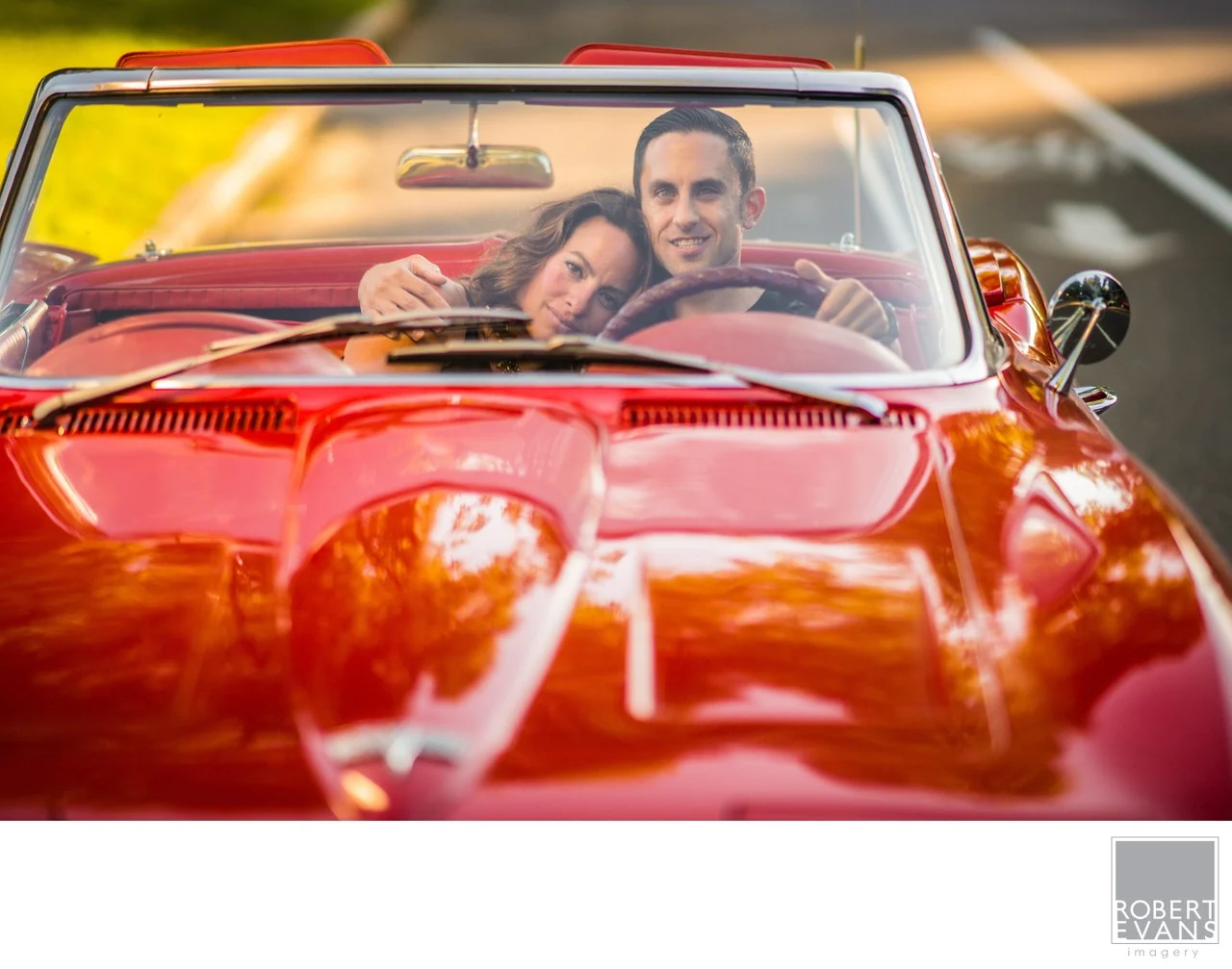 A couple leaning together in a red convertible, with warm light across the hood.