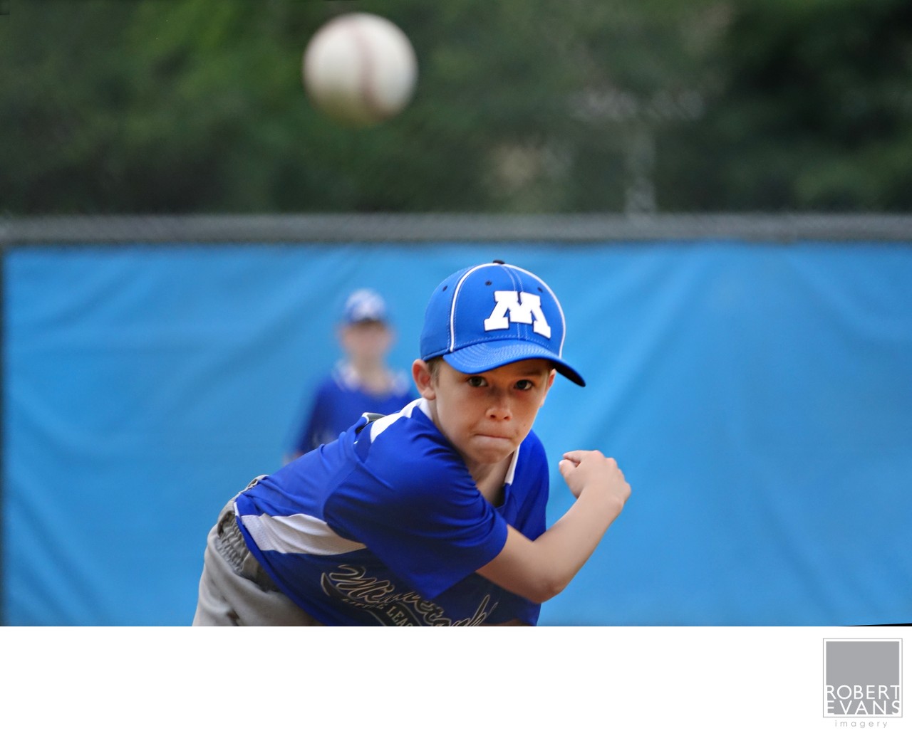 A young baseball pitcher in blue follows his throw with the ball blurred in the foreground.
