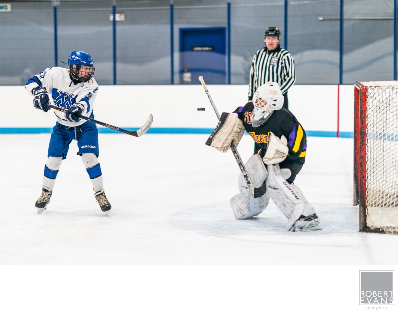 A youth hockey player prepares a shot in front of a goalie and official.