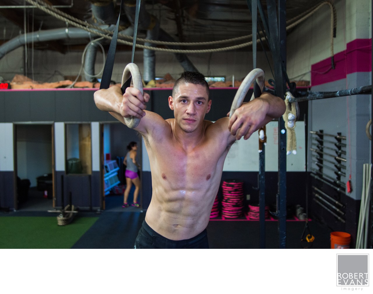 A shirtless athlete supports his weight on rings inside a training gym.