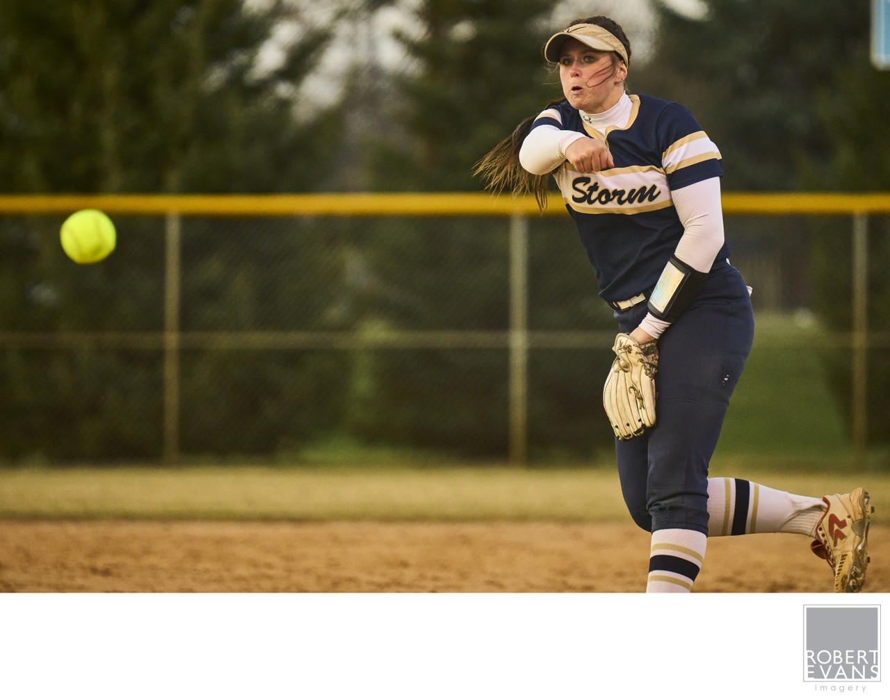 A pitcher in a Storm uniform follows through as a yellow softball travels toward the batter.