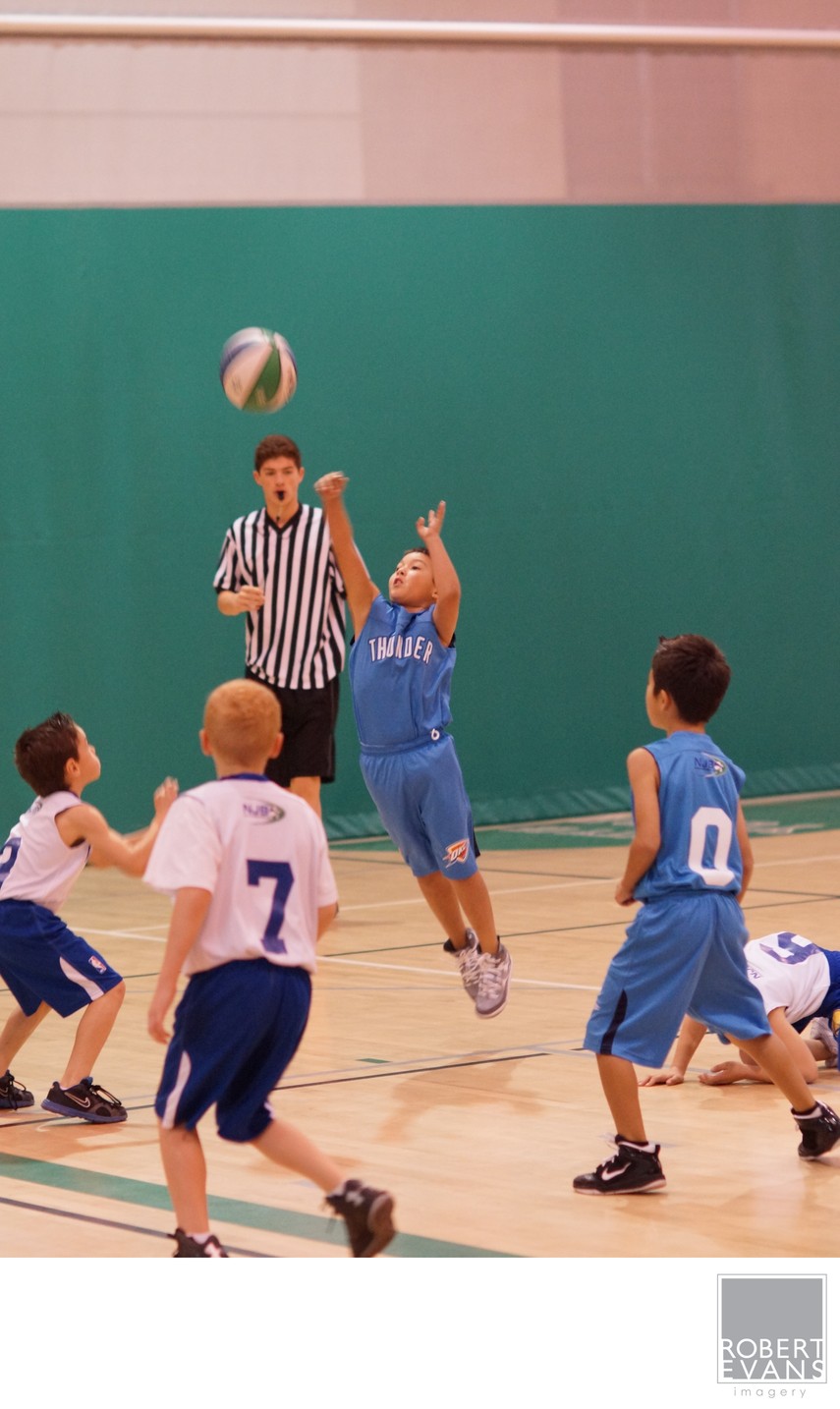 Two young basketball players reach for the ball as an official tosses it upward.