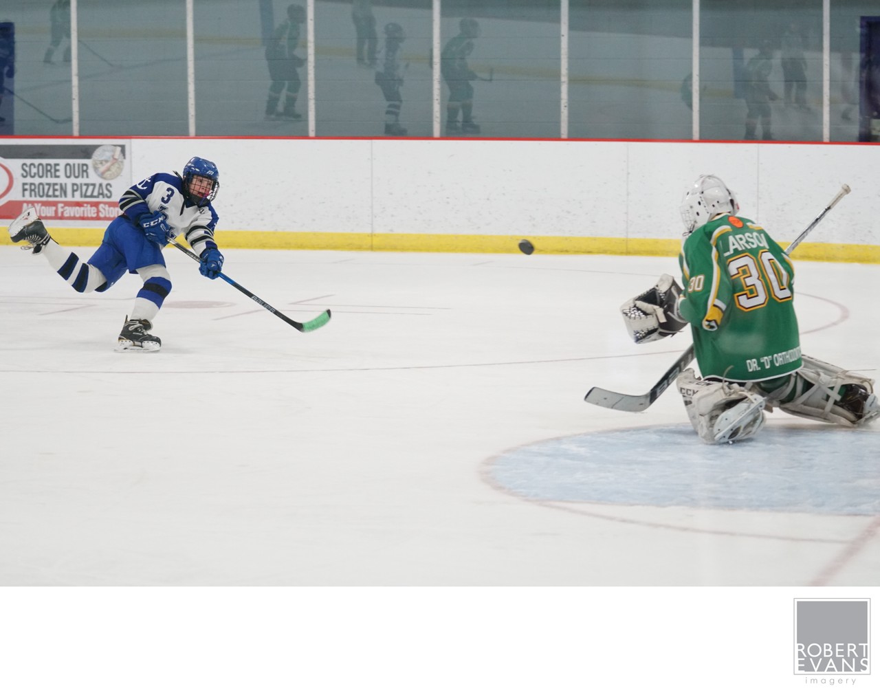 A blue-and-white attacker skates toward a green-uniformed goalie with open ice between them.