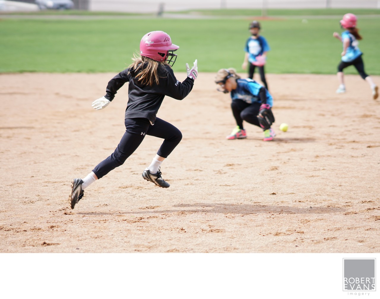 A player wearing a pink helmet runs across the dirt as a fielder crouches behind her.