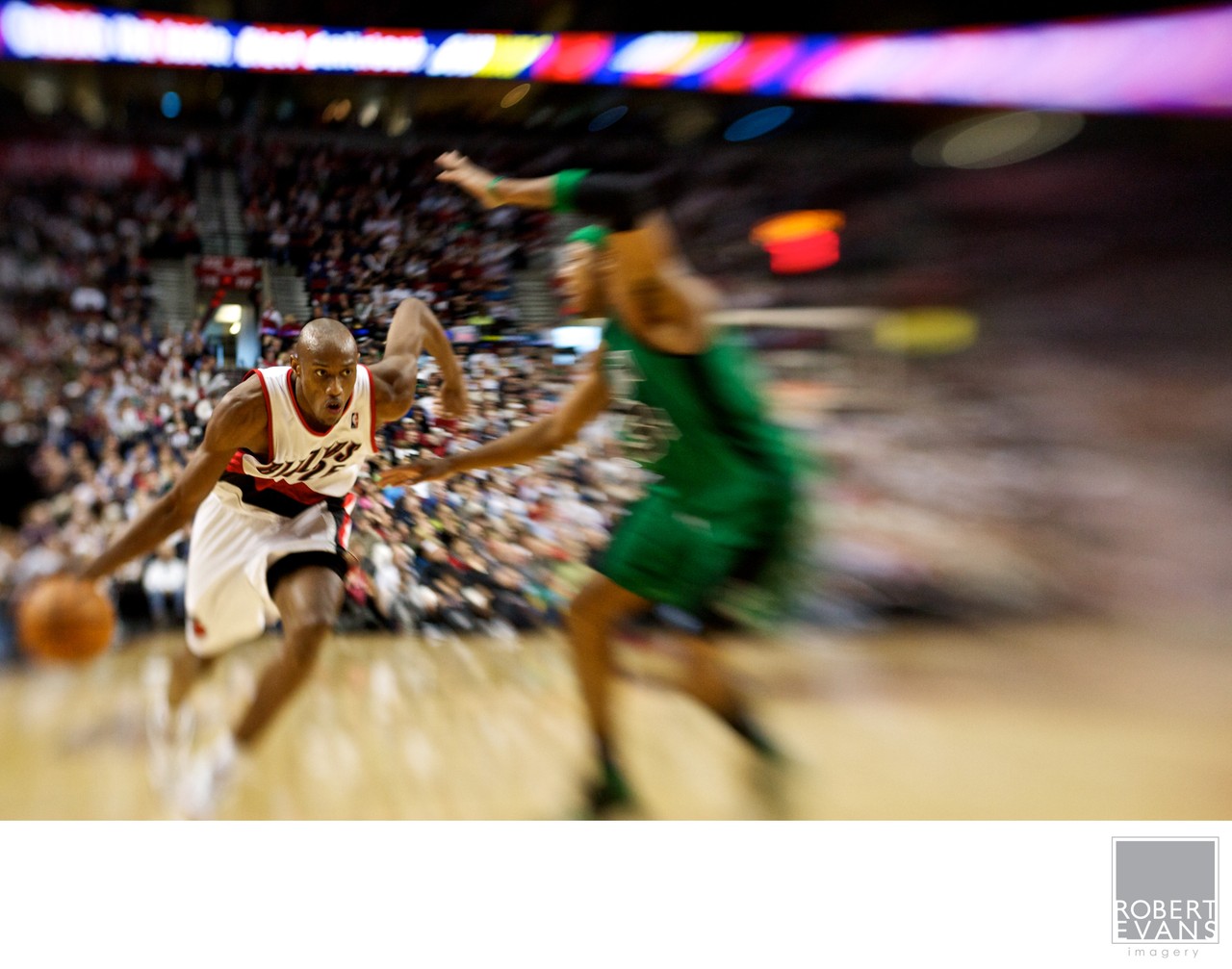 A basketball player in white moves forward with the ball as a green-clad defender follows.