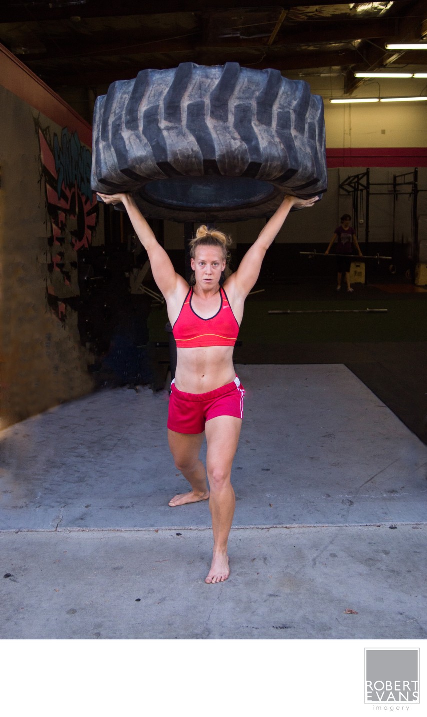 A woman in training clothes holds a large tire above her head outside a gym.
