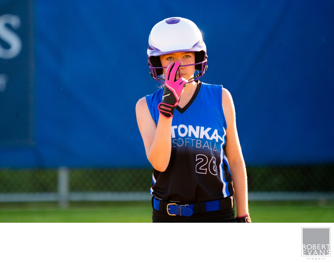 A young softball player in a blue uniform adjusts her bright pink face guard.
