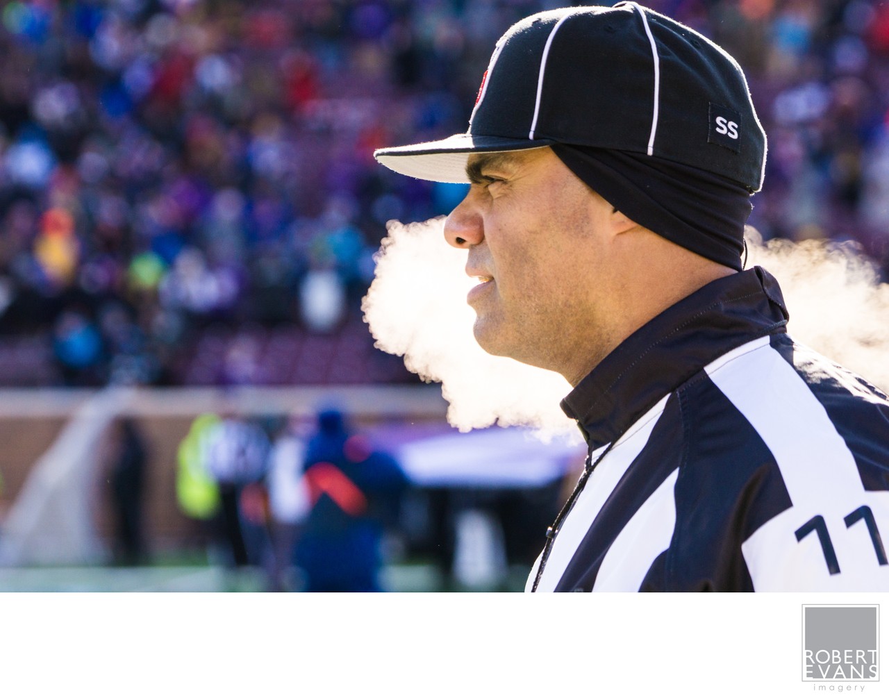 A football official exhales visible breath on a cold day beside the field.