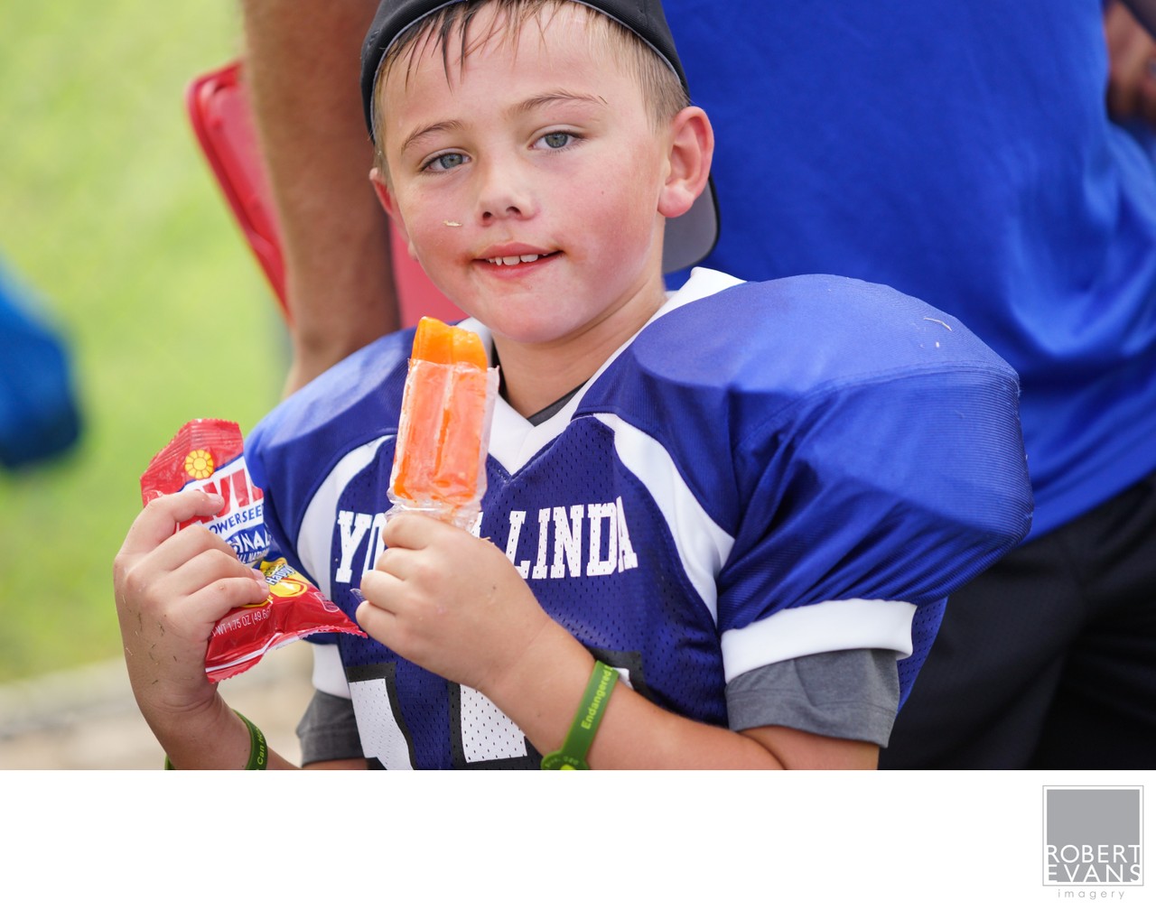 A young player in blue shoulder pads holds a colorful frozen treat beside the field.