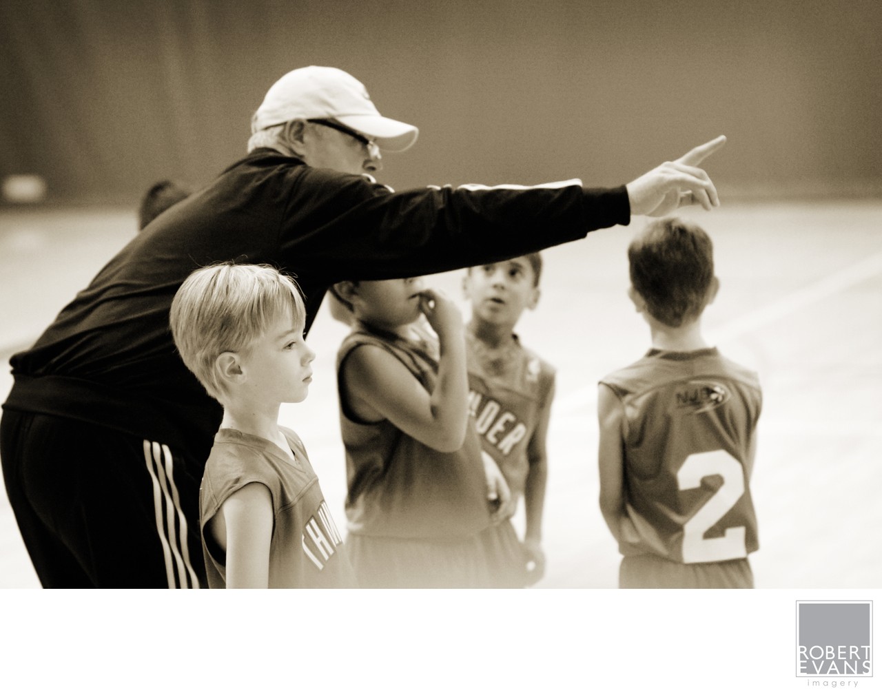 A coach points across the court while children listen nearby in monochrome.