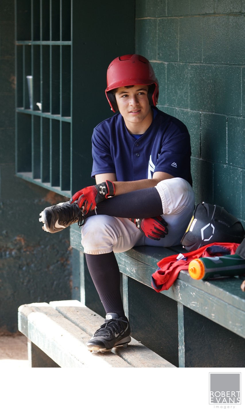 A baseball player wearing a red cap sits alone with his glove on a dugout bench.