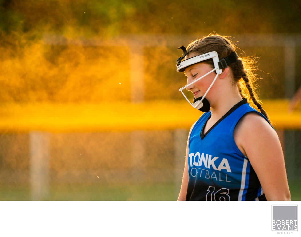 A player in a blue uniform and face guard looks downward in golden light.