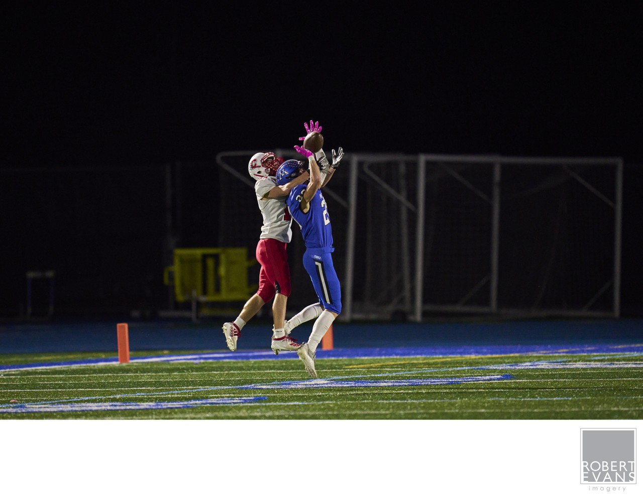 Two football players leap and reach for a pass under stadium lights.