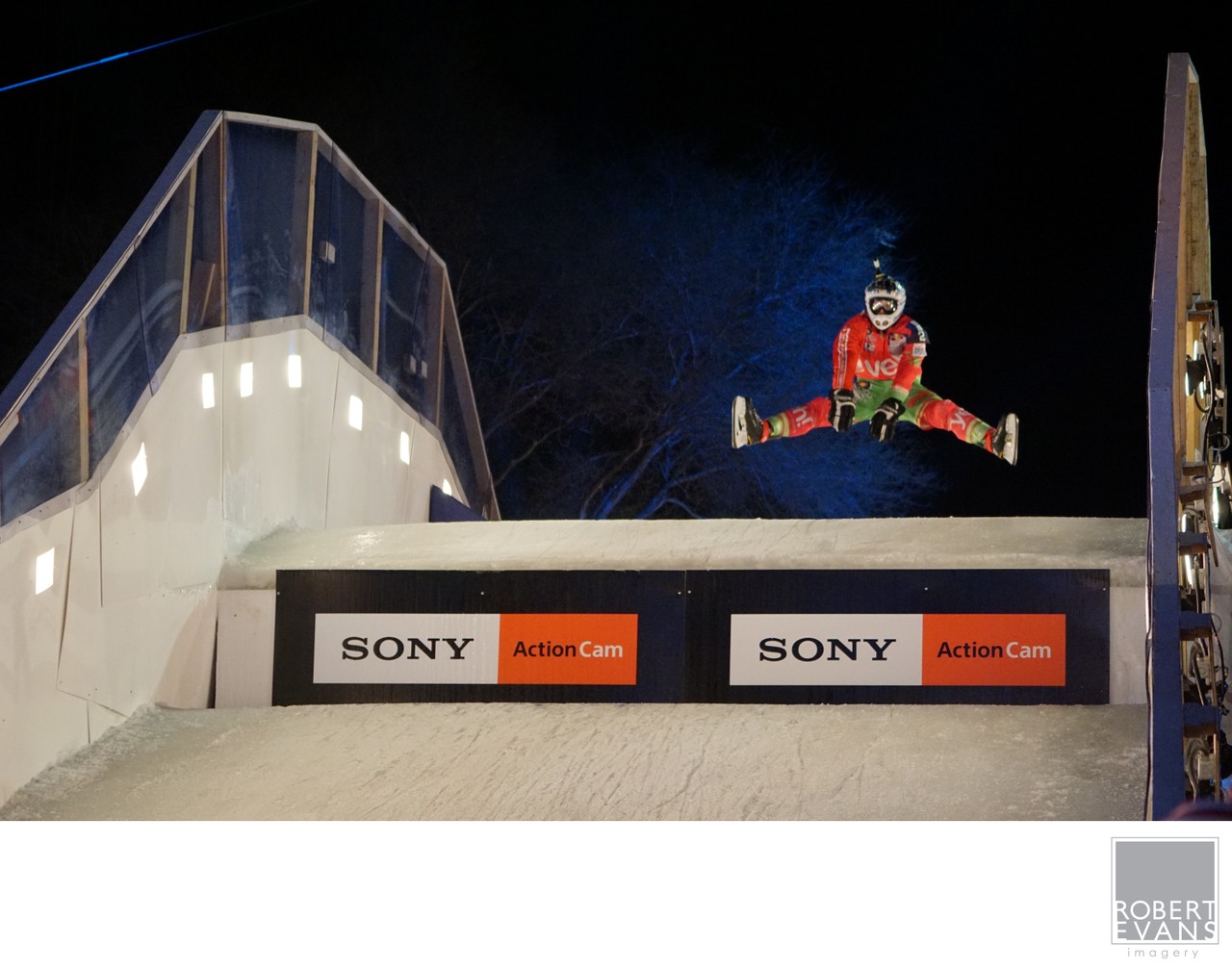 A racer in protective equipment jumps high above an illuminated ice course at night.