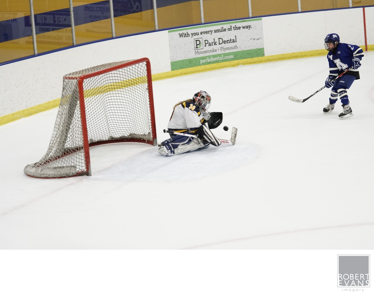 A hockey goalie stretches toward a low puck while an attacker closes in.
