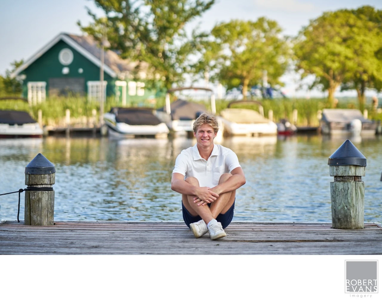 A young man in a white shirt sits cross-legged on a dock with boats and a waterfront building behind him.