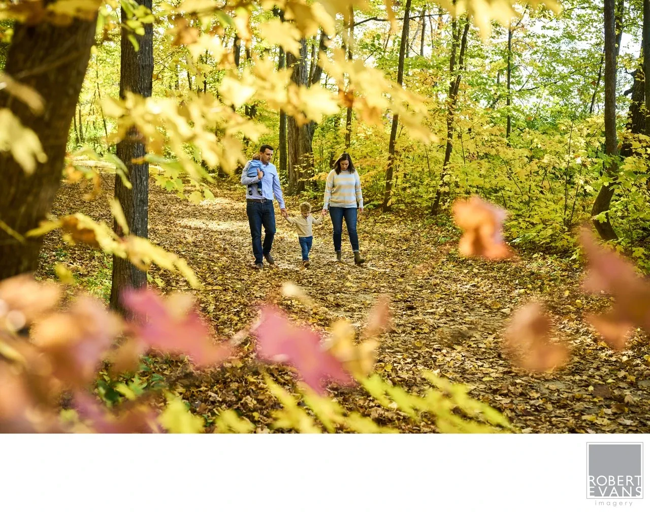 Two adults and a small child walk away along a path framed by colorful autumn leaves.