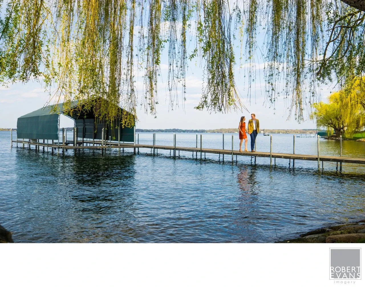 Two people stand together on a long lakeside walkway beneath overhanging willow branches.