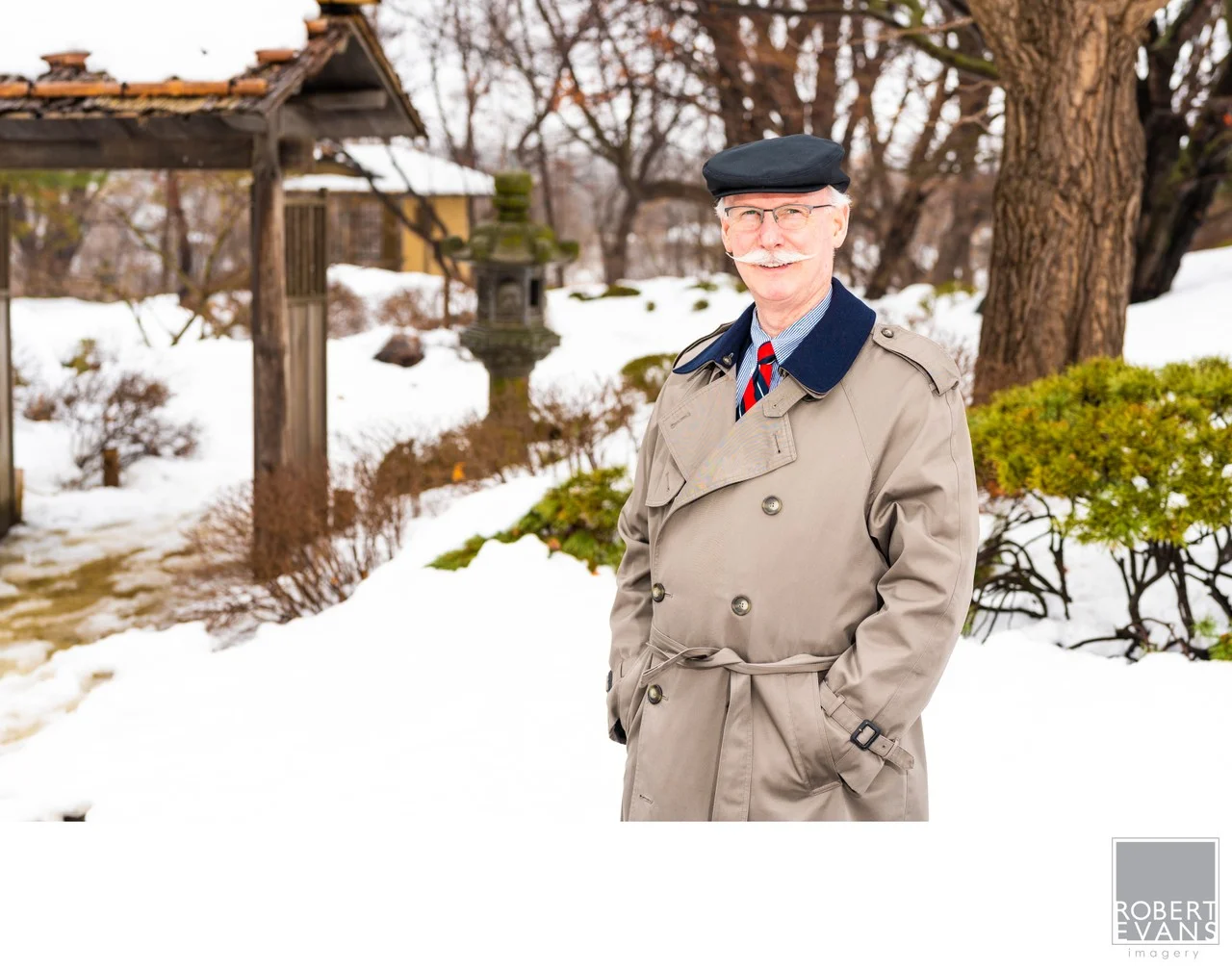 An older man in a hat and trench coat stands beside a tree in a snow-covered garden.