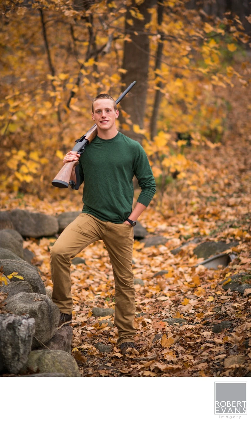 A young man in a green sweater stands with one foot on a rock beneath golden autumn trees.