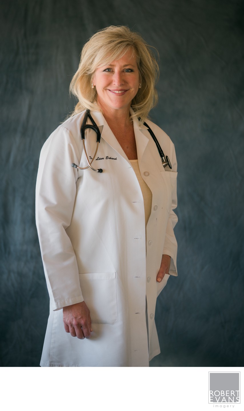A smiling woman wears a white medical coat and stethoscope against a softly lit studio background.