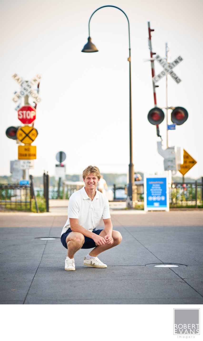A young man crouches on a paved sidewalk beneath railroad crossing signs.