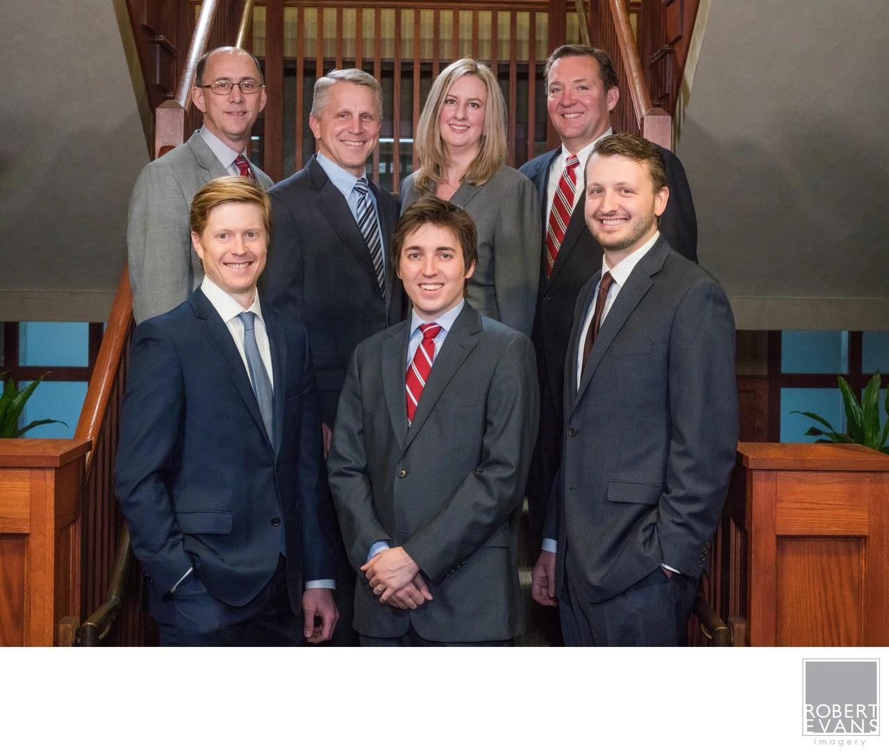 A group of seven adults in formal clothing poses together beside an indoor staircase.