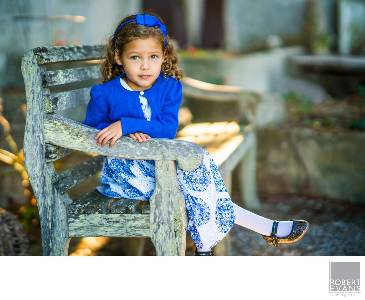 A small girl wearing blue sits on a weathered wooden bench outdoors.