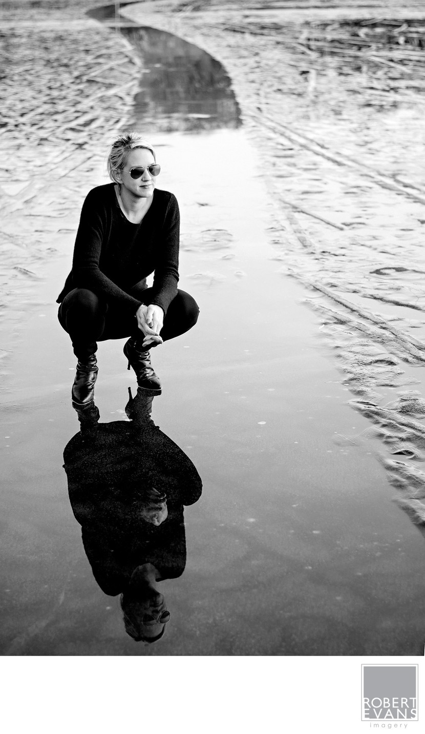 A person crouches beside a long puddle, creating a mirrored black-and-white portrait.