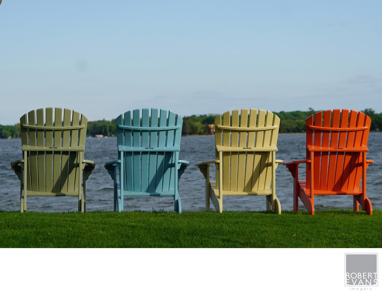 Green, blue, yellow and red Adirondack chairs face the water from a grassy shore.