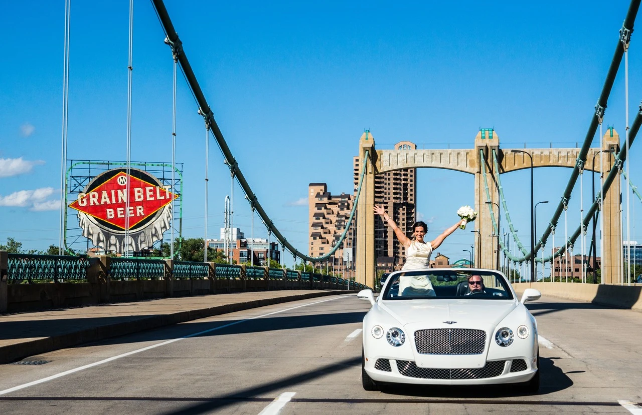 A bride stands in a white convertible on a suspension bridge under a blue sky.