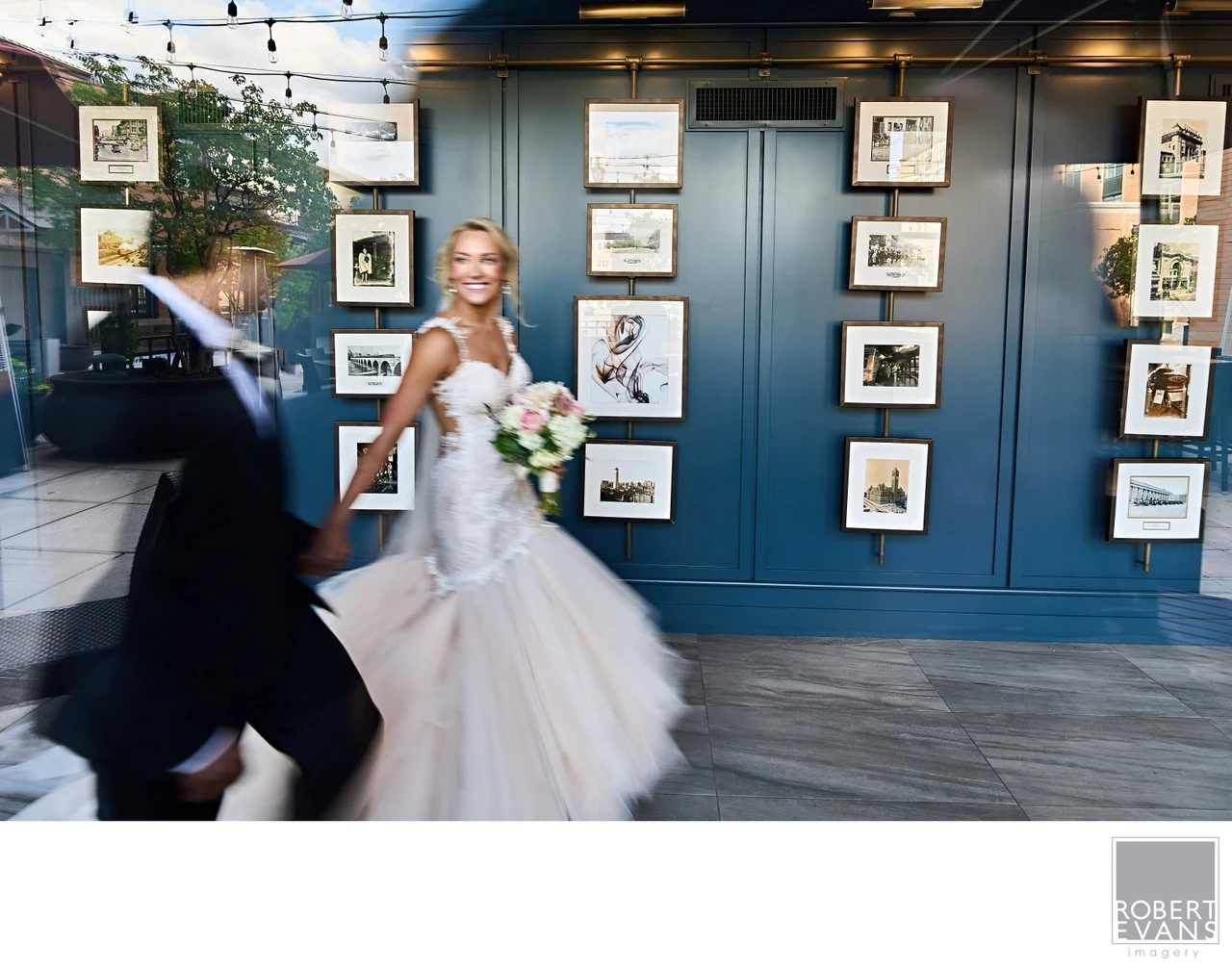 A bride smiles as she moves with her groom past framed artwork indoors.