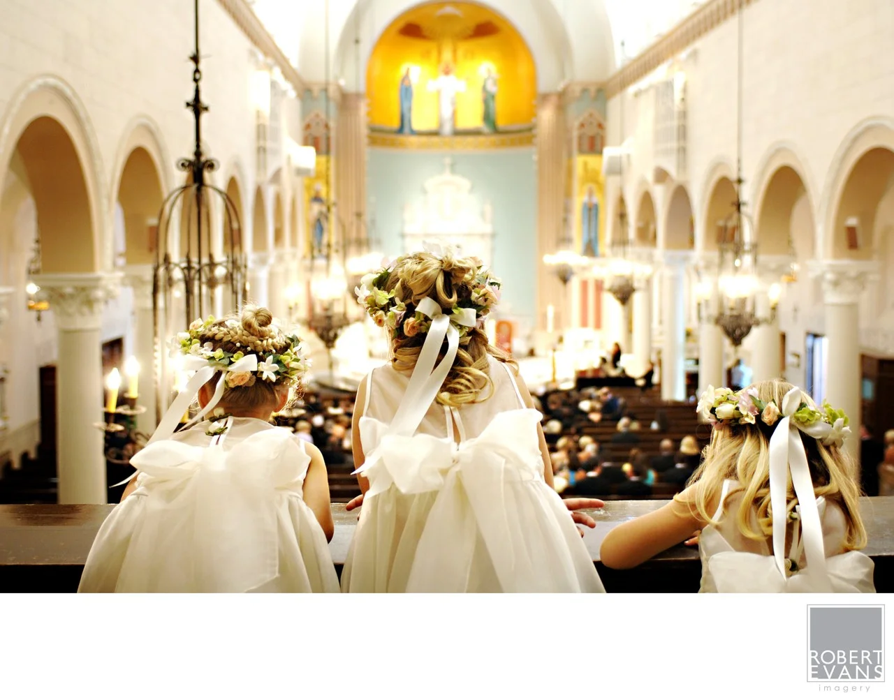 Flower girls in white dresses face the altar inside a warmly lit church.
