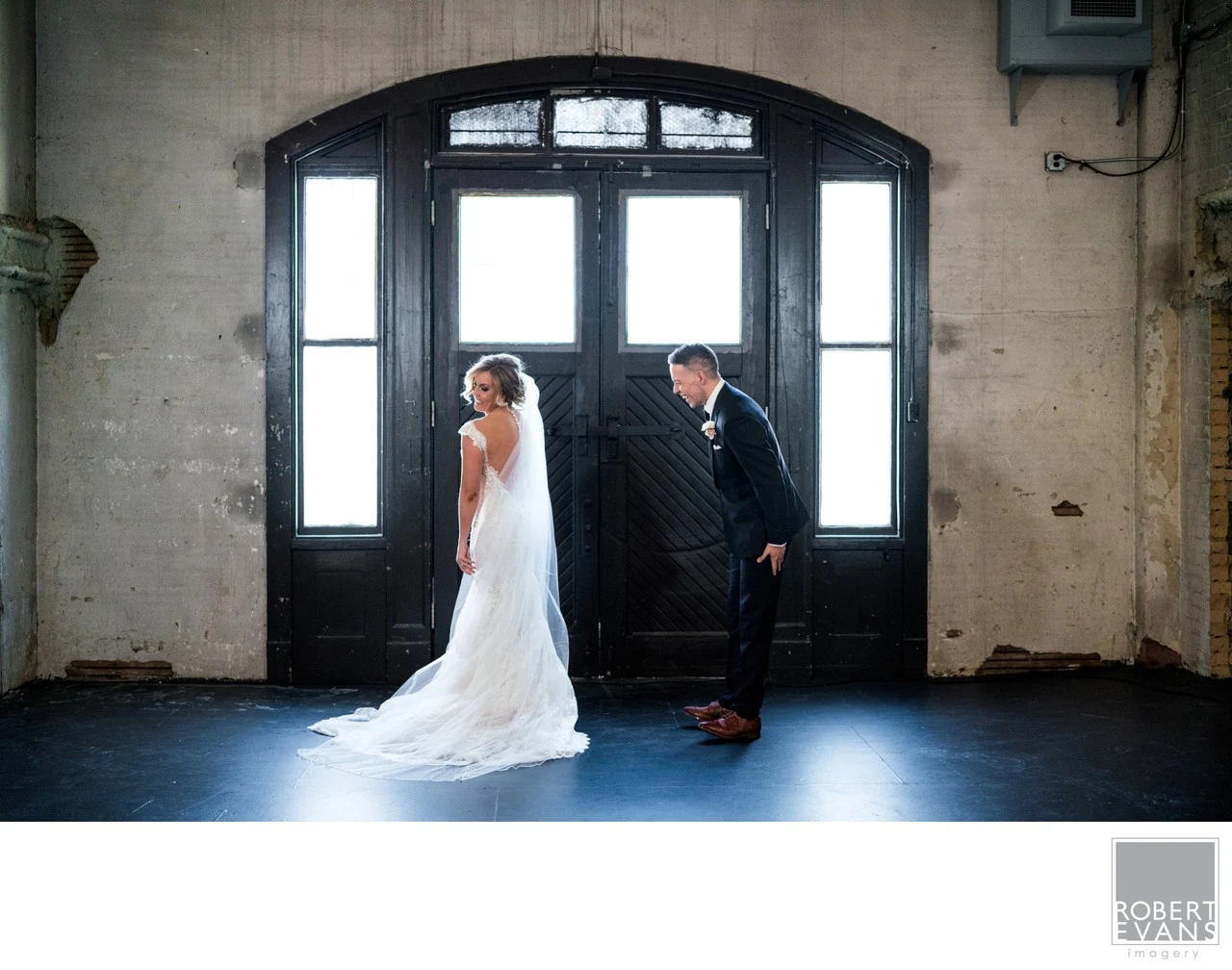 A bride and groom stand apart beneath tall arched black doors in an industrial interior.