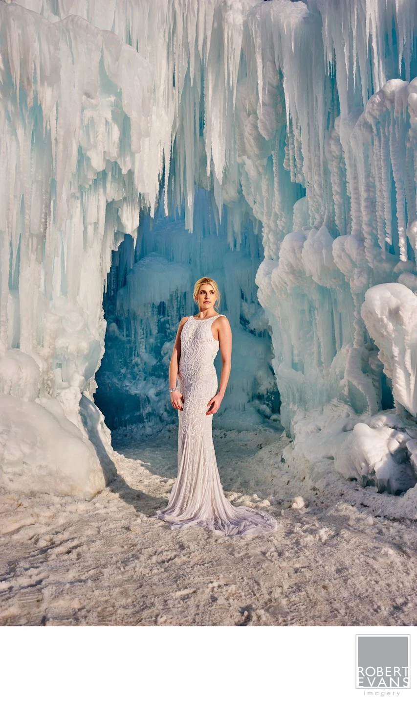 A bride in a fitted gown stands inside towering pale blue ice formations.