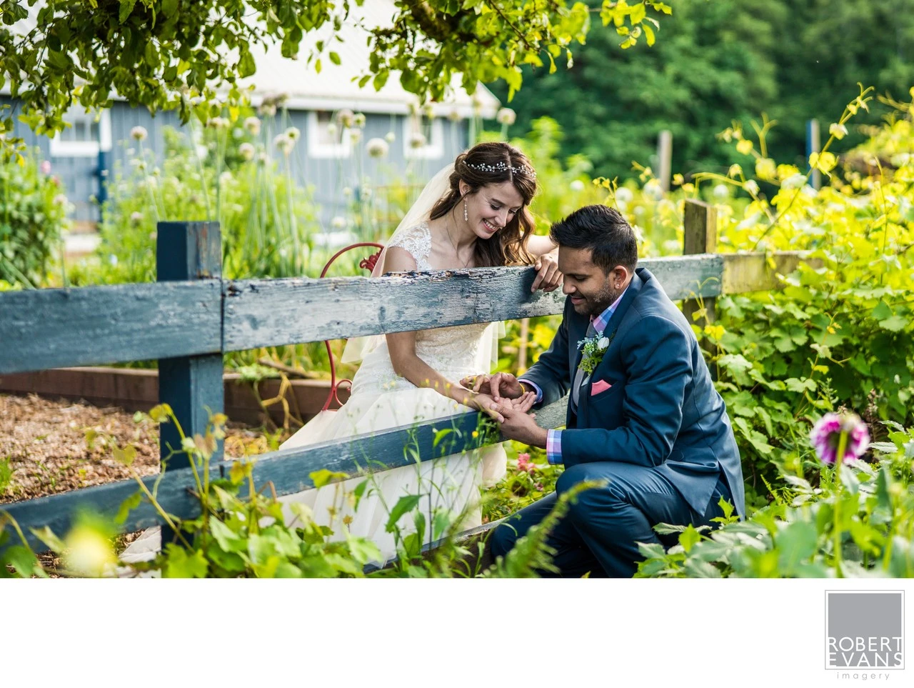 A bride and groom sit together beside a rustic fence surrounded by green plants.