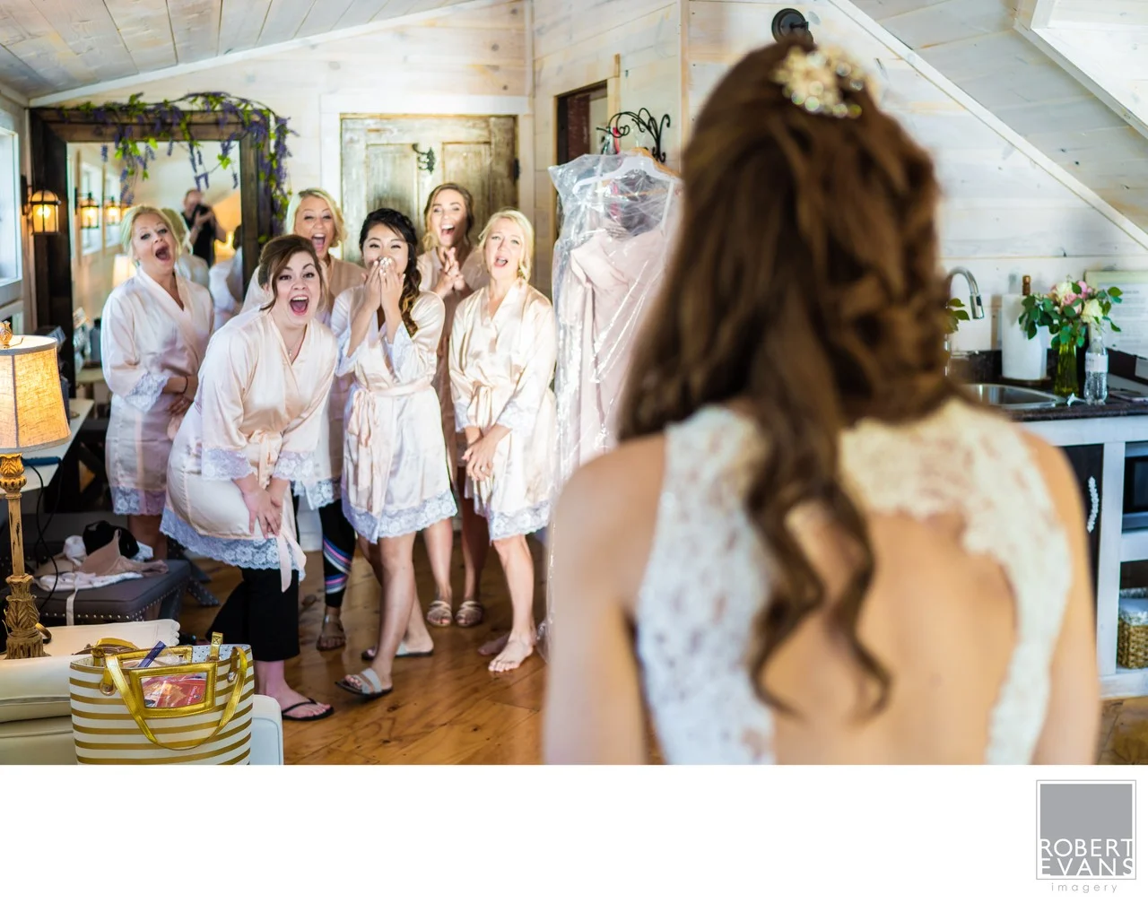 Bridesmaids react with delighted expressions as a bride enters the room in her wedding gown.