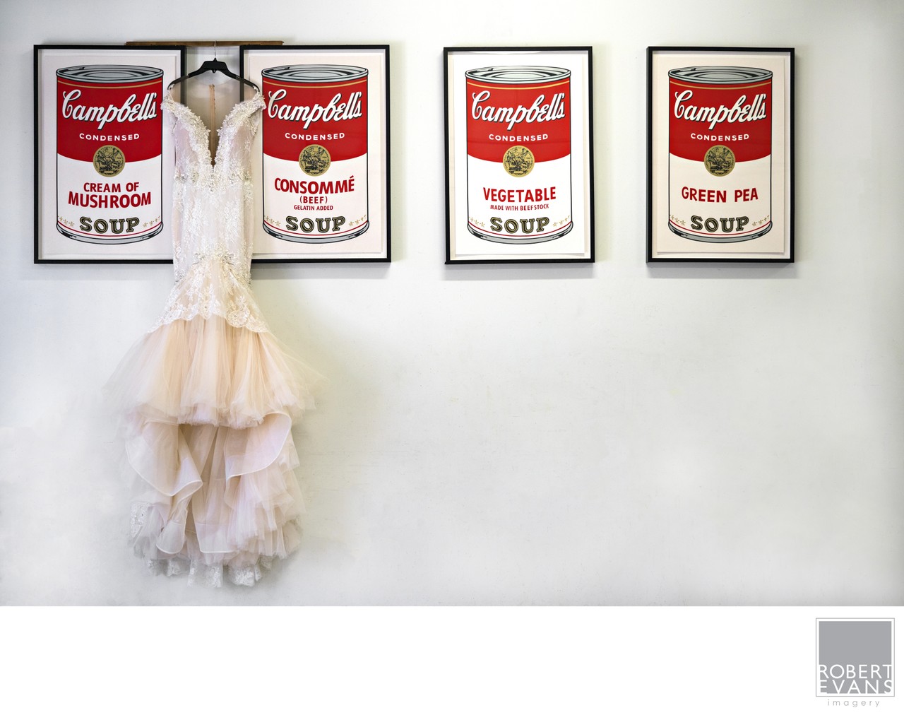A wedding dress hangs beneath a row of framed soup-can artworks.